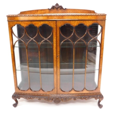 An early 20thC walnut and burr walnut bow fronted display cabinet, with a raised back above a leaf carved frieze and two astragal glazed doors, enclosing glass shelves on shell carved short cabriole legs with pad feet, 133cm high, 116cm wide.