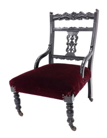 An Edwardian ebonised nursing chair, with a carved and pierced back, a red upholstered padded seat, on turned legs with ceramic castors.