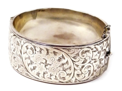 A silver bangle, with engraved floral and foliate decoration, on a snap clasp, with safety chain as fitted, 1.1oz.