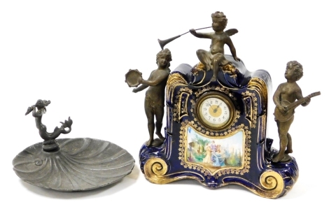 A late 19thC porcelain cased mantel clock, circular dial bearing Arabic numerals, the cobalt blue case with reserve panel decorated with 17thC pastoral figures in a garden, surmounted by spelter putti playing musical instruments, 29cm high, together with 