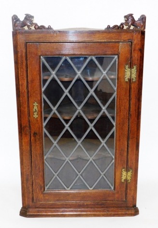 An early 20thC oak hanging corner cupboard, the top carved with two lions, the glazed door enclosing three serpentine shaped shelves, raised on a plinth base, 109cm high, 65cm wide, 29cm deep.