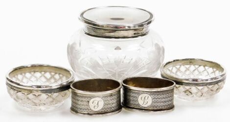 A George V cut glass toilet jar with silver lid, Birmingham 1918, 1oz, a pair of oval silver napkin rings, initialled engraved, indistinct Birmingham hallmarks, 0.80oz and a pair of cut glass salts with silver rims, London 1916.