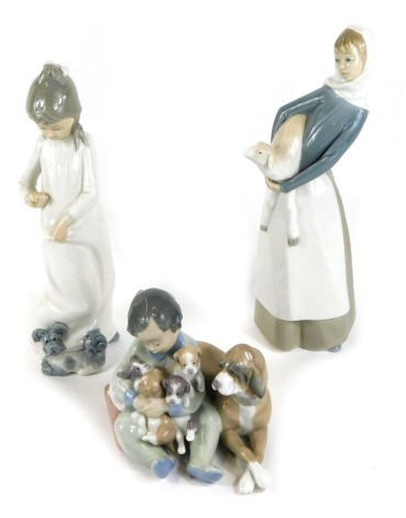 A Lladro porcelain figure group of a seated boy holding a clutch of puppies, a dog by his side, a further Lladro figure of a standing lady holding a lamb, together with a Nao figure of a girl with a puppy pulling at her skirt tails. (3)