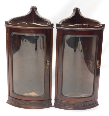 A pair of 20thC mahogany hanging corner cupboards, each with a blind fret above glazed doors, with further scrolls and a floral moulding, 72cm high, 39 wide, 32cm deep.