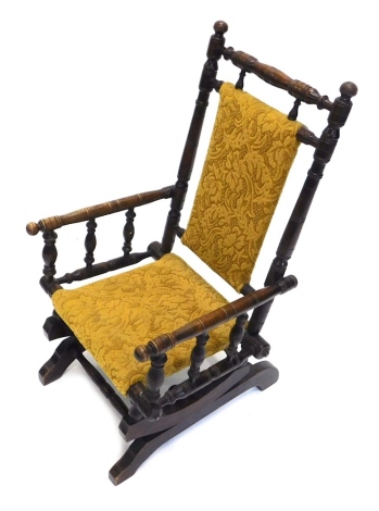 An early 20thC walnut child's American rocking chair, overstuffed to the back and seat in later floral gilt material, with turned back and arms, on scroll feet, 82cm high.