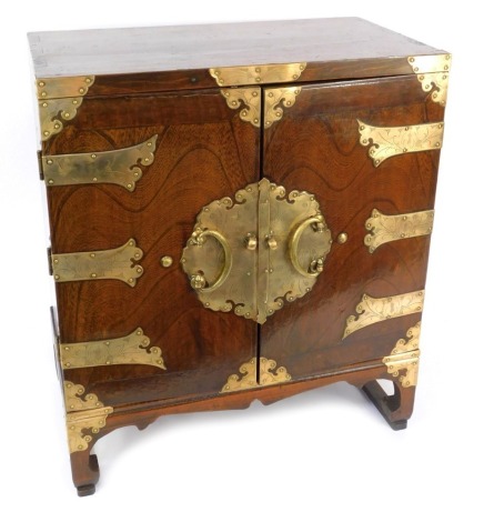 An early 20thC Korean elm and brass bound table cabinet, the two doors opening to reveal a shelf above a pair of small frieze drawers, raised on inward scroll feet united by side stretchers, 63cm high, 55cm wide, 31cm deep.