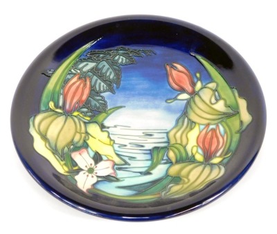 A Moorcroft Pottery plate decorated with pond flowers, against a blue background, painted and impressed marks, 26cm wide, boxed.