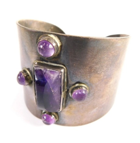 A Mexican torc bangle, with applied cabochon and carved rough cut amethyst central plate, on a white metal cuff stamped sterling Mexico, 6cm diameter, 79.6g all in.