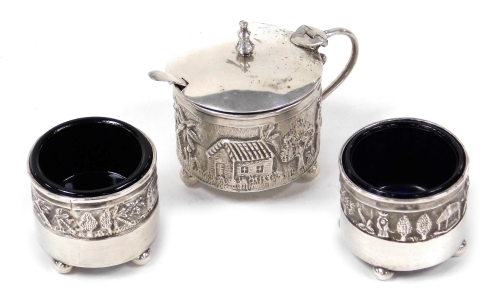 A 20thC Indian silver three piece cruet, comprising lidded mustard pot and two open salts, each piece embossed with figures and buildings in a rural setting, blue glass liners, later hallmarked, Henry Charles Freeman and J.B. & Co., Birmingham 1911 and 19