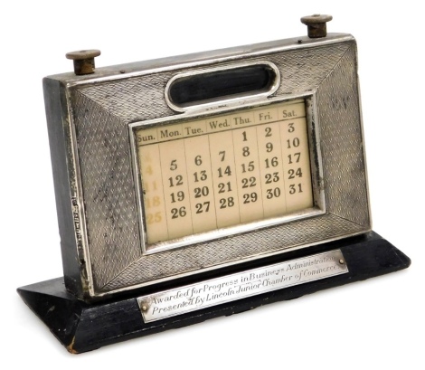 An Elizabeth II silver faced desk calendar, engine turned, with paper central label and twin dials, mounted on a black painted base bearing presentation plaque Awarded For Progress In Business Administration Presented By Lincoln Junior Chamber of Commerce