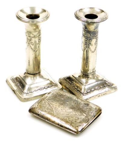 A pair of Edward VII silver squat candlesticks, embossed with bows, swags, flowers, etc., each on a silver base, loaded, Henry Matthews, Birmingham 1905, 13cm high (AF), together with a silver cigarette case, bright cut decorated with flowers and scrolls,