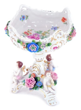 A Sitzendorf porcelain tazza, the basket with four moulded handles, printed with reserves of flowers, and encrusted with flowers, with applied cherubs and flower to the tripod base, printed marks, 20cm high, 16cm wide.