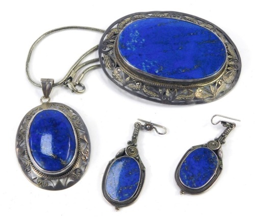 A lapis lazuli jewellery suite, comprising large oval brooch, with pierced border, white metal stamped 925, 8cm x 6cm, a similar design pendant, 6.5cm high, on modern chain, 40cm long, and a pair of matched drop earrings, white metal unmarked 5cm high, 89