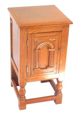 An oak bedside cupboard, with an arched carved panelled door opening to reveal a single shelf, raised on turned supports united by a box stretcher, 63cm high, 34cm wide, 33cm deep.