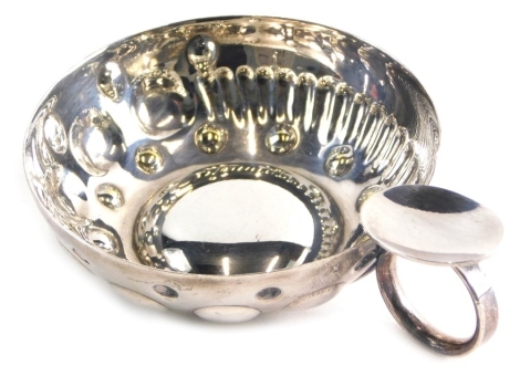 A silver coloured testavine or wine taster, with circular body, hand chased bead and a swirl fluted decoration, with loop handle and plain thumb piece, on a concave base, x makers mark near handle, 4cm high.