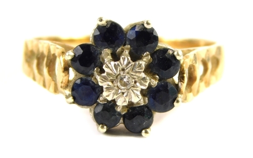 A 9ct gold dress ring, with floral cluster set with central tiny diamond in illusion setting, surrounded by eight sapphires, in a raised basket, with pierced design and hammered shoulders, ring size N½, 3.3g all in.