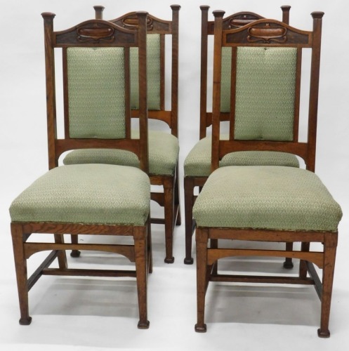 A set of four oak Arts and Crafts dining chairs, each with high backs, stylised carved lotus flower arched tops, overstuffed backs and seats in later green material, on front square legs terminating in compressed square feet, 109cm high.