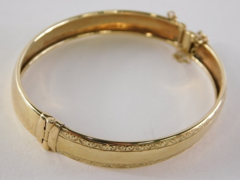 A 9ct gold hinged bangle, of half inch design with engraved borders, clasp and safety chain, 6cm diameter, yellow metal unmarked, 9.1g.