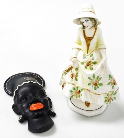 A Katzhutte pottery figure of a lady, modelled standing holding her dress and a fan, raised on an octagonal base, printed mark, 27cm high, and a 1950s black pottery face mask wall plaque of a lady, 20cm high. (2)