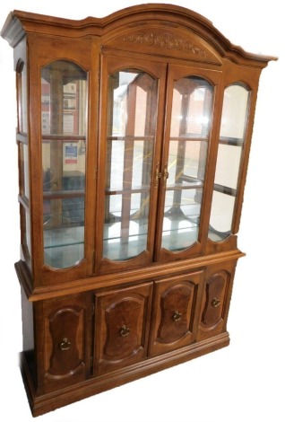 A 20thC mahogany display cabinet, the arched top with a scroll carved panel above two glazed doors, enclosing two shelves with a mirrored back, above two doors enclosing a sectioned tray and a shelf, on a plinth base, 206cm high, 144cm wide, 41.5cm deep.