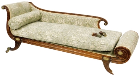 An Empire style oak framed chaise longue, upholstered in green fabric decorated with birds and flowers, raised on scroll legs, brass capped on castors, 211cm wide, (AF).