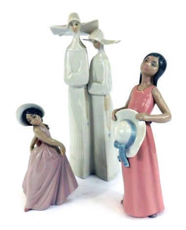 Three Lladro figures, comprising a figure group of two nuns, in white matt finish, 34cm high, a figure of a girl in pink dress with hat, 25cm high, and a figure of a young child in pink dress, 20cm high. (3)