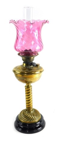 An early 20thC brass oil lamp, with cranberry glass shade, plain glass chimney, and raised brass reservoir, on turned stem and inverted circular foot, 71cm high.