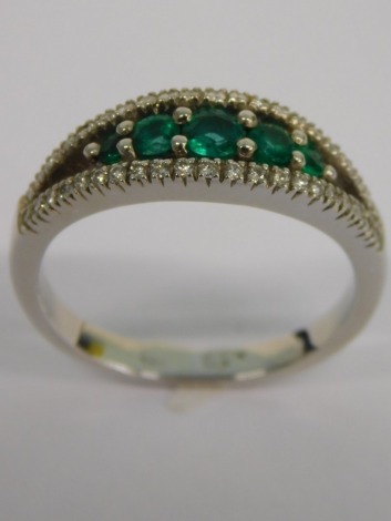 An emerald and diamond dress ring, the centre set with five round brilliant cut emeralds in claw setting, with V splayed diamond shoulders totalling approx 0.19ct, on a white metal band, unmarked believed to be 18ct, 4.6g all in.