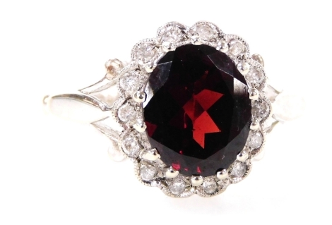 A 9ct white gold, garnet and diamond cluster ring, with a central oval cut garnet in multi claw setting, with petallated border set with tiny diamonds, with V splayed shoulders, ring size O, 3.7g all in.