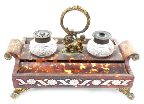 A Regency tortoiseshell desk stand, with gilt serpentine handle above a cherub and recumbent lion, having two plain scroll handles to the ends and incorporating two pen rests and a cut glass pounce jar and matching ink receiver, with borders of flower hea