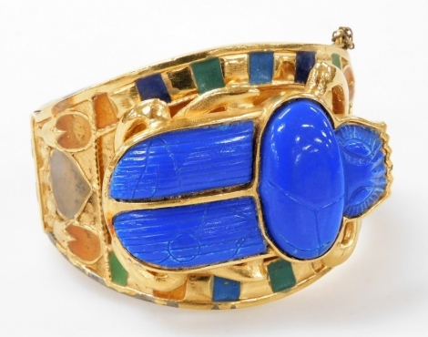 A late 20thC Egyptian revival scarab bangle, by Thomas Fattorini Limited, with an imitation carved lapis lazuli scarab beetle, within enamel foliate and geometric banding, in a yellow metal hinged setting, with safety chain.