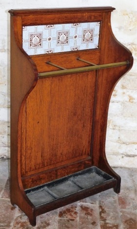 A late Victorian oak umbrella stand, the panelled back inset with Minton style ceramic tiles, decorated with pomegranates in Morris style, with a brass rail and a triple drip tray, on shaped end supports, 101cm high, 52cm wide.