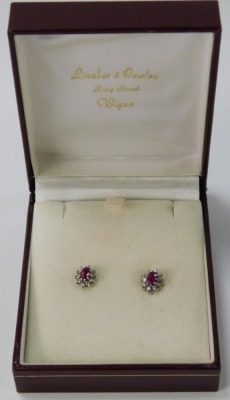 A pair of 9ct white gold ruby and diamond earrings, each formed as a cluster with central oval cut ruby surrounded by ten round brilliant cut tiny diamonds, each in a white claw setting, on single pin back with matched different backs, 2g all in, boxed. - 2