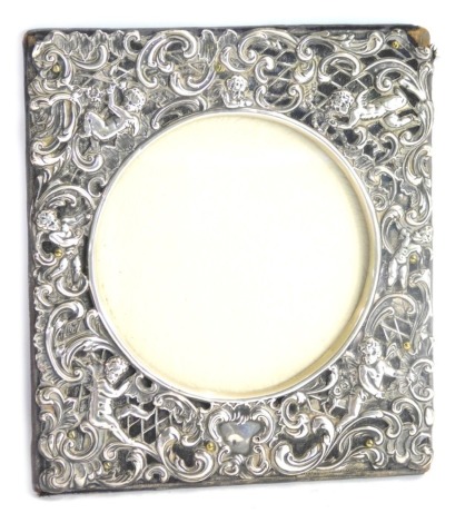 An Edward VII silver strut photograph frame, embossed with putti and rococo scrolls, vacant shield reserve, strut lacking, William Comyns, London 1904, aperture 10cm wide, frame 16cm x 14.5cm.