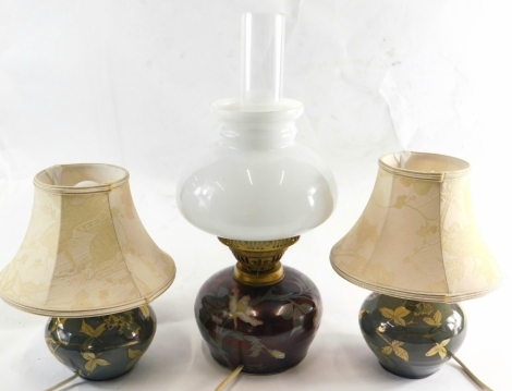 Three Jonathan Chiswell Jones studio pottery lustre lamps, comprising a pair of table lamps in green and an oil lamp in red lustre, decorated with flowers, with clear chimney and bulbous body, 65cm high. (3) Buyer Note: WARNING! This lot contains untested