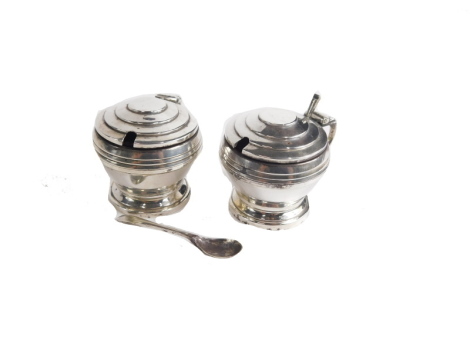 A pair of George V silver Art Deco mustard pots, by Walker and Hall, each with stepped circular lids, scroll handles and circular feet, Sheffield 1934, with associated spoons, 6cm high, 5oz.