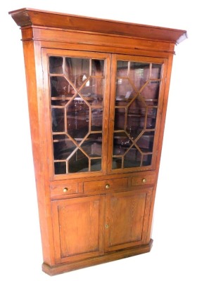 A pine standing corner cabinet, with a moulded cornice, above two astragal glazed doors enclosing shaped shelves, the base with a drawer flanked by two false drawers, above two panelled doors, on a plinth, 187cm high, 115cm wide.