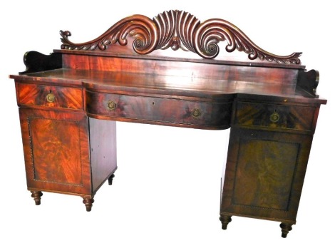 A George IV mahogany sideboard, the raised back carved with scrolls, flanking a central shelf, the base with a central bowed drawer, flanked by two further frieze drawers, above two doors with beaded panels, on turned legs, 41cm high, 182cm wide.