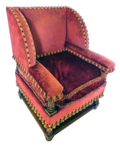 An oak framed wingback chair, upholstered in red and gold velvet, with tasselled borders.