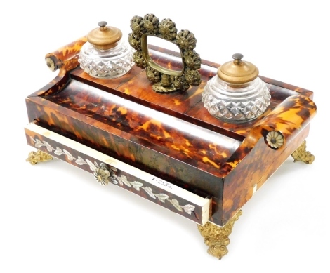 A 19thC French tortoiseshell encrier, with scrolling side handles, and brass carrying handle, having two cut glass central inkwells, flanked by pen recesses, above a frieze drawer with mother of pearl inlay, raised on floral embossed scrolling feet, 25cm 