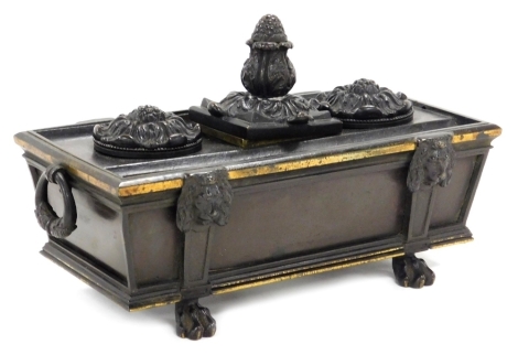 A late 19thC bronze desk stand, formed as a sarcophagus, with twin ring handles, the top with a central pineapple finial enclosing a glass inkwell, flanked by two floral lids enclosing a pounce pot and an inkwell, raised on paw feet, 21cm wide.