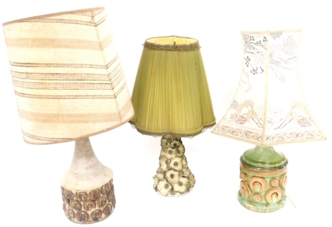 Three mid century pottery table lamps, comprising a floral encrusted lamp with a green frilled shade, 49cm high, green glazed cylindrical pottery lamp, with moulded circular decoration, with an octagonal floral shade, 53cm high, and a buff stoneware potte