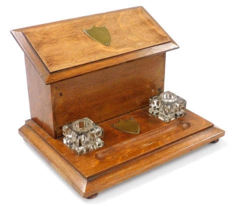 A Victorian oak desk stand, with brass shield mounts, having a lidded waterfall stationary box, behind two cut glass inkwells and a pen recess, raised on a plinth base, on turned feet, 20cm high, 30cm wide, 23cm deep.