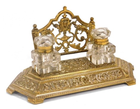 A Victorian brass desk stand, with embossed foliate scroll decoration, the top having two cut glass inkwells with hinged cut glass lids, 30cm wide.