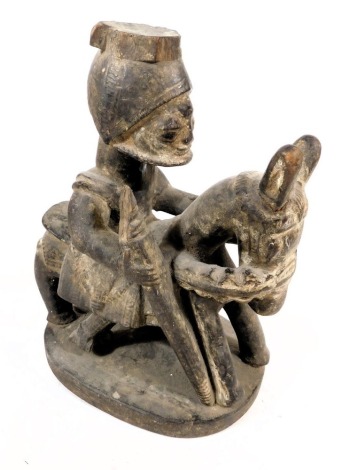 A late 19thC West African wooden carving, of a man in a headdress, mounted on a horse or donkey, and holding a staff, raised on an oval base, traces of white pigment, 21cm wide.