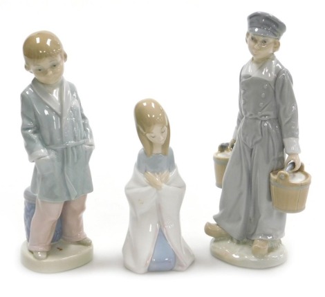 Three Lladro porcelain figures, comprising a boy in a dressing gown with a stool behind, Dutch boy holding two milk pails (AF), and a kneeling figure of a girl. (3)