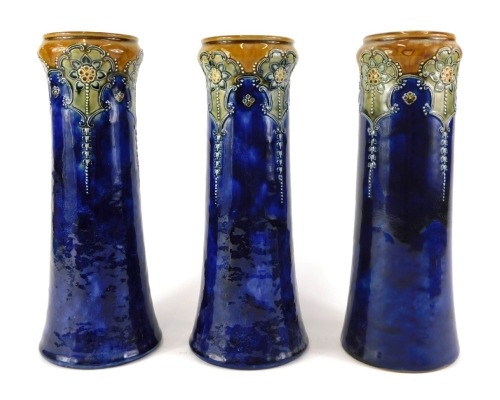 Three Royal Doulton stoneware vases, each decorated with flowers, stylised leaves in Art Nouveau style, in green, on a mottled blue ground, impressed marks to underside, indistinct makers monograms, each 32cm high.