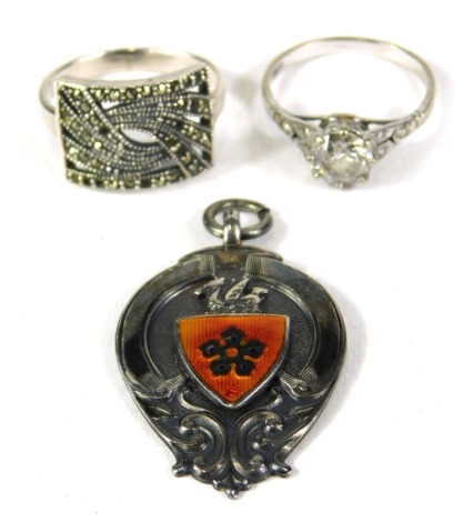 A group of silver costume jewellery, comprising a silver and marcasite dress ring, a silver and paste stone set solitaire ring, and a silver and enamel crest, with orange shield, 13.9g all in. (3)