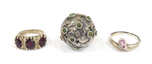 Three dress rings, comprising a white metal bore ring set with green and other rubbed agate, with faint hallmarks, a silver and red paste stone set dress ring, and an unmarked plated purple paste stone set dress ring. (3)