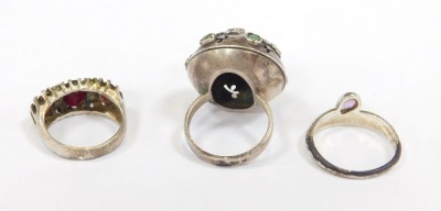 Three dress rings, comprising a white metal bore ring set with green and other rubbed agate, with faint hallmarks, a silver and red paste stone set dress ring, and an unmarked plated purple paste stone set dress ring. (3) - 2